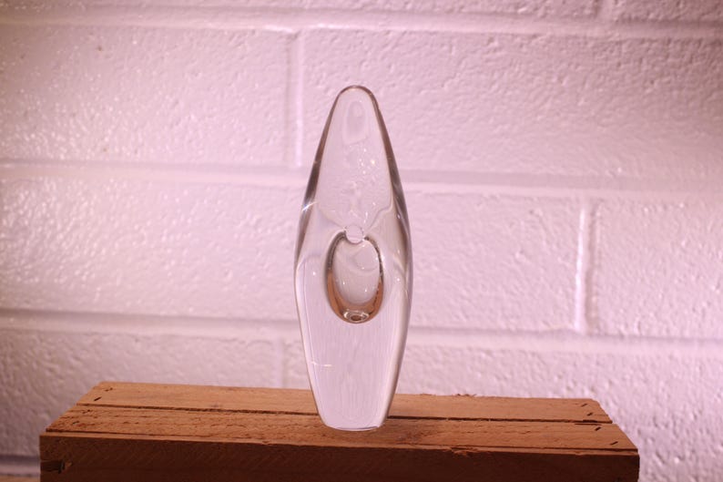 May include: A clear glass vase with an elongated, oval shape and a smaller, circular bubble detail. The vase is displayed on a wooden surface against a white brick wall. The vase is approximately 10 inches tall.