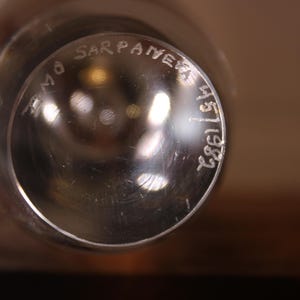 May include: Close-up of a clear glass object with the text "Timo Sarpaneva 45/1982" etched on the surface. The glass has a reflective quality, with blurred light reflections visible.