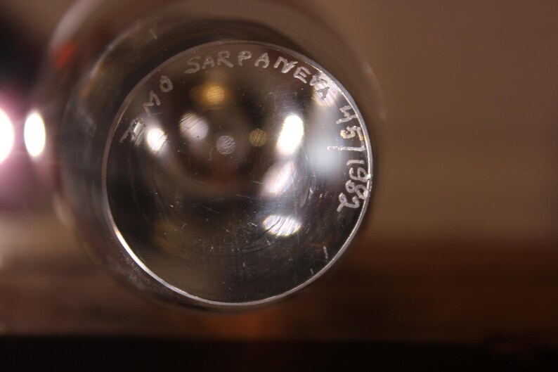 May include: Close-up of a clear glass object with the text "Timo Sarpaneva 45/1982" etched on the surface. The glass has a reflective quality, with blurred light reflections visible.