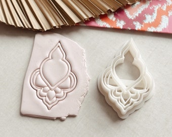 May include: A white plastic cookie cutter in the shape of a decorative, ornate, teardrop-shaped design. The cutter is shown next to a pink impression of the cutter in a block of modeling clay.