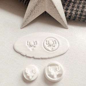 May include: Four white plastic cookie cutters in the shape of a deer head. Two of the cutters are round and two are square. The cutters are on a white surface.