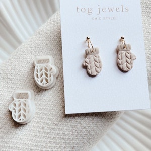 May include: A pair of beige knitted mitten earrings with gold hoops. The earrings are displayed on a white card with the text "tog jewels chic style".