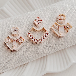 May include: Three beige earring cutters with floral and geometric patterns are displayed on a white, textured surface. The cutters are arranged on a white, scalloped-edge plate, suggesting a craft or jewellery-making theme.