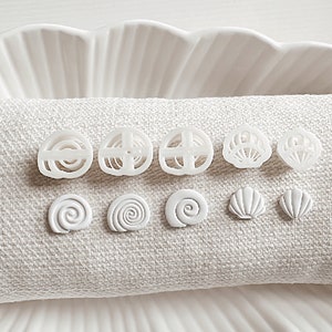 Peut inclure: Un ensemble de boucles d'oreilles en céramique blanche avec différents motifs inspirés de la mer, notamment des coquillages, des spirales et des motifs géométriques.
