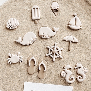 May include: A collection of white ceramic jewelry charms with a beach theme. The charms include a seashell, a whale, a crab, a sailboat, an ice cream cone, a popsicle, flip flops, a steering wheel, an umbrella, and two seahorses. The charms are arranged on a light brown surface that resembles sand. The text "tog jewels chic style" is printed on a white card.