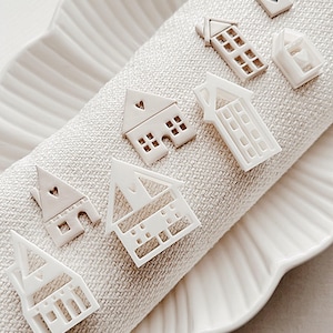 May include: A set of seven white ceramic house-shaped ornaments with a heart detail on each. The ornaments are arranged on a white fabric surface.