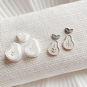 May include: A set of four pear-shaped earrings. Two earrings are white with a leaf design and two earrings are a light brown with a leaf design.