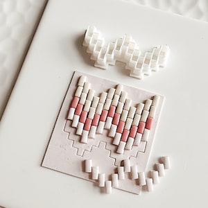 May include: A white tile with a pink and white pixelated design. The design is a heart shape with a white outline and pink interior. There are extra white tiles scattered around the design.