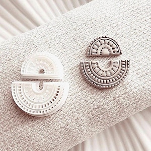 May include: Two half-circle shaped polymer clay earring cutters, one white and one beige, with intricate geometric patterns. The cutters are placed on a textured, off-white fabric, suggesting a craft or DIY project. The design is reminiscent of a sun or a stylized arch.