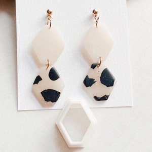 May include: A pair of dangle earrings with two geometric shapes. The top shape is a light pink, and the bottom shape is a white and black geometric pattern.