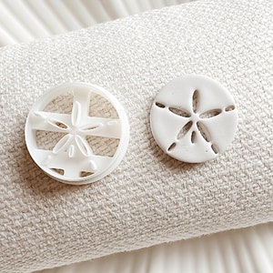 May include: Two white plastic cookie cutters shaped like a sand dollar. The cutter on the left is open and the cutter on the right is closed.