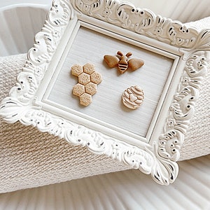 Peut inclure: Un cadre blanc orné avec un fond brun clair. À l'intérieur du cadre, trois petits objets décoratifs : un nid d'abeilles, une abeille et une ruche. Les objets sont faits d'un matériau brun clair.