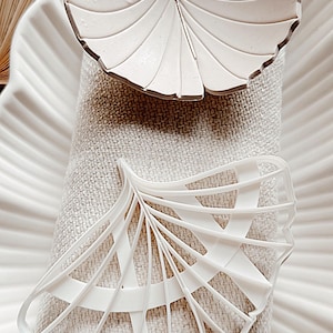 May include: Two white ginkgo leaf cookie cutters, one solid and one with a lattice design. The cutters are on a white linen background.