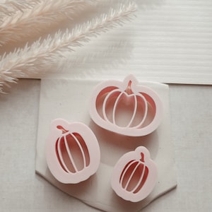 May include: Three pink plastic pumpkin cookie cutters of different sizes. The largest cutter is a traditional pumpkin shape with a stem. The other two cutters are more elongated and have a stem and a curved bottom.