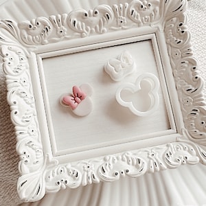 May include: Three white plastic cookie cutters in a white frame. The cutters are shaped like a crown, a mouse head, and a mouse head with a pink bow.