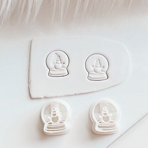 May include: Two white plastic cookie cutters shaped like snow globes with a snowman inside. The cutters are on a white surface. Two more cutters are shown in the background, one on each side of a white rectangular piece of clay.