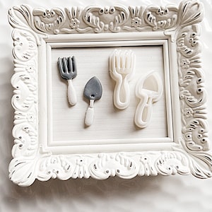 May include: Four white plastic cookie cutters in the shape of gardening tools: a fork, a trowel, and two different styles of rakes. The cutters are displayed in a white ornate frame.