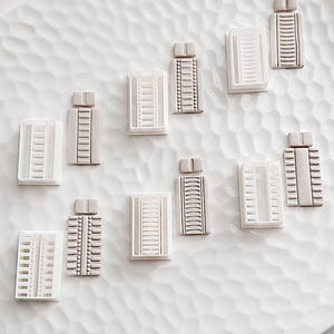 Peut inclure: Dix petites décorations de bâtiments en céramique blanche et brune disposées sur une surface texturée blanche. Les ornements sont en forme de bâtiments hauts avec des fenêtres et des détails.