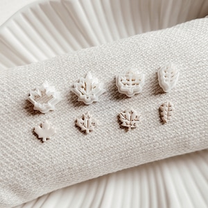 May include: A set of seven white plastic leaf-shaped cookie cutters on a white fabric surface. The cutters are arranged in a row, with the largest cutter on the left and the smallest cutter on the right.