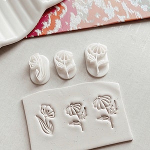 May include: A set of six white polymer clay stamps featuring different flower designs. The stamps are arranged in two rows on a white surface. The top row has three stamps, each with a different flower design. The bottom row has three stamps, each with a different flower design.