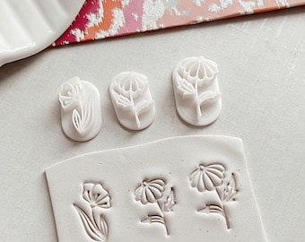 May include: A set of six white polymer clay stamps featuring different flower designs. The stamps are arranged in two rows on a white surface. The top row has three stamps, each with a different flower design. The bottom row has three stamps, each with a different flower design.