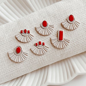 May include: A set of six white ceramic earrings with red geometric shapes. The earrings are displayed on a white fabric surface.