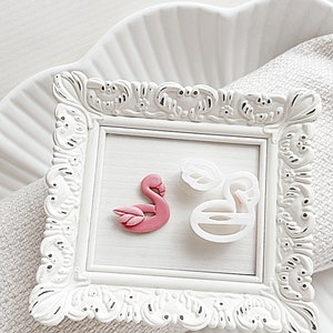 May include: A white frame with a pink flamingo and a white flamingo cookie cutter inside. The frame is ornate and has a white background.