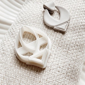 May include: Two white ceramic earrings, one shaped like a leaf and the other shaped like a curved abstract design. The earrings are on a white fabric background.