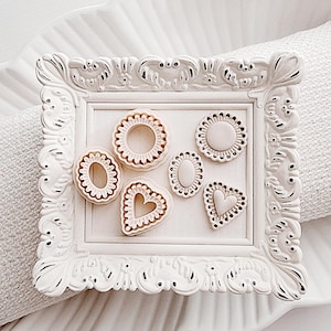 May include: A white, ornate picture frame holds six decorative, off-white shapes with a scalloped edge and a heart shape. The frame and shapes are on a white, scalloped plate.