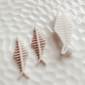 May include: Three white ceramic fish skeletons on a white textured surface. The fish skeletons are detailed with ribs and a tail fin.