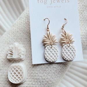 Peut inclure: Boucles d'oreilles en céramique blanche en forme d'ananas avec des crochets dorés. Les boucles d'oreilles sont sur une carte blanche avec le texte "tog jewels chic style".