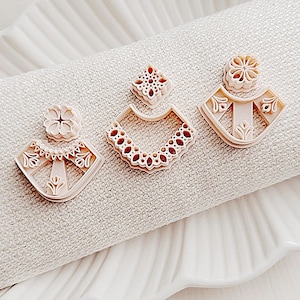 May include: Three beige polymer clay earring cutters in various shapes, including a flower, a square, and a fan-like design with intricate details. The cutters are displayed on a white, textured surface.