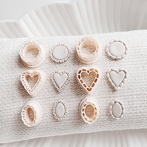 May include: A collection of light pink cookie cutters in various shapes, including circles, ovals, and hearts, with decorative edges. The cutters are arranged on a white, textured surface, and displayed on a white, scalloped dish.