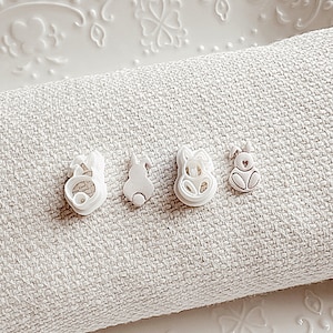 May include: Four white ceramic bunny cookie cutters on a white linen surface. The cookie cutters are in a row and are all facing the same direction.