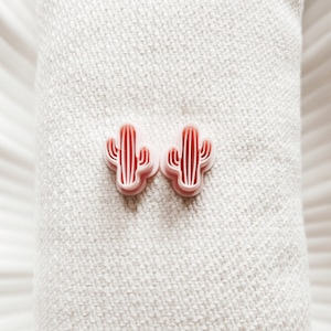 Peut inclure: Deux boucles d'oreilles en forme de cactus roses sur un fond de tissu blanc.