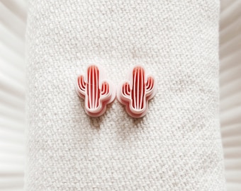 Peut inclure: Deux boucles d'oreilles en forme de cactus roses sur un fond de tissu blanc.