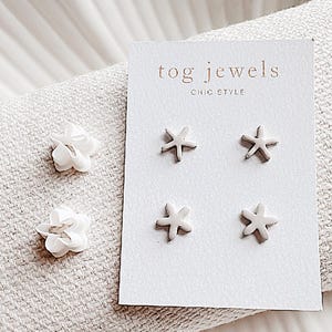 May include: A set of four white ceramic earrings. Two earrings are shaped like a flower and two are shaped like a star. The earrings are displayed on a white card with the text "tog.jewels chic style".