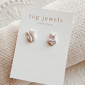 May include: A pair of silver cat-shaped earrings on a white card with the text "tog jewels chic style".