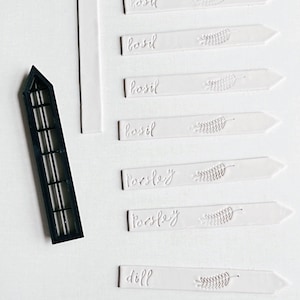 May include: White plant markers with the words "basil", "parsley", and "dill" written on them. The markers are arranged in a row on a white surface.