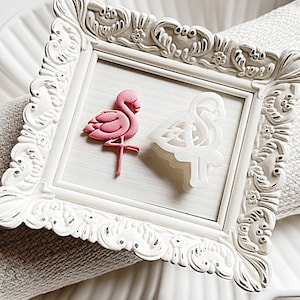 May include: A white ornate picture frame with a pink flamingo and a white flamingo cutout inside. The frame is sitting on a white surface.