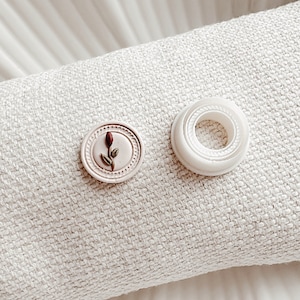 May include: Two round buttons, one white and one pink with a small brown flower design. The buttons are on a white fabric surface.