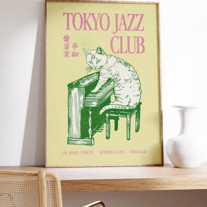 May include: A framed poster with a light yellow background features a green illustration of a cat playing a piano. The text "TOKYO JAZZ CLUB" is in pink. Japanese text and address details are also present.