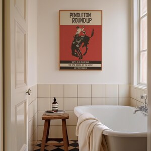 May include: A framed vintage poster with the text "PENDLETON ROUND UP" and a rodeo scene graphic hangs above a white bathtub. The poster has a red background and is in a bathroom setting with a wooden stool and a bottle.