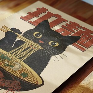 May include: A black cat with big eyes is eating noodles from a bowl with chopsticks. The cat is looking directly at the viewer. The background is a light brown with the Japanese word "Ramen" in red.