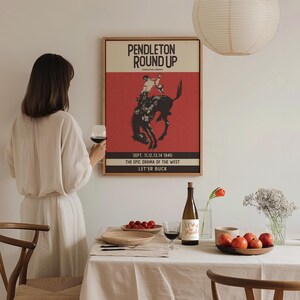 May include: A framed vintage poster with the text "PENDLETON ROUND UP" and a rodeo illustration. The poster is mounted on a white wall. A table is set with a white tablecloth, wine glasses, and a bottle of wine. The text "SEPT. 11.12.13.14 1940 THE EPIC DRAMA OF THE WEST LET'ER BUCK" is also visible.