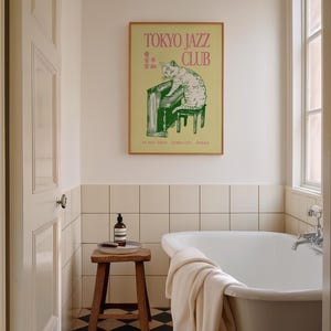 May include: A framed poster with the text "TOKYO JAZZ CLUB" in pink lettering. The poster features a green illustration of a cat playing a piano. The poster is hanging above a white bathtub in a bathroom setting.
