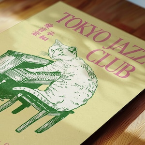 May include: A light yellow poster with the text "TOKYO JAZZ CLUB" in pink. A white cat is illustrated playing a green piano. Japanese text is also present. The poster is on a wooden surface.