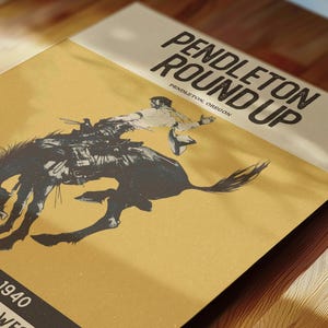May include: A vintage poster with the text "PENDLETON ROUND UP" and "PENDLETON, OREGON" in bold, black font. The poster features a black and white illustration of a rodeo cowboy on a bucking bronco. The background is a tan color.