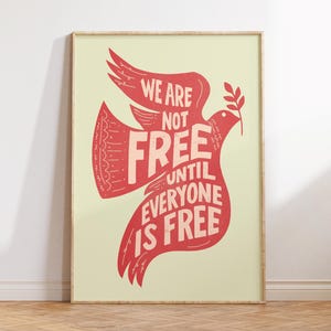 May include: A framed art print with a red dove design holding an olive branch. The dove contains the text "WE ARE NOT FREE UNTIL EVERYONE IS FREE". The background is pale yellow, and the frame is a light wood colour. The print is displayed on a wooden floor.
