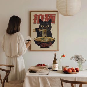 May include: A black cat with a bowl of noodles in front of it. The cat is holding chopsticks and eating the noodles. The image is in a Japanese style and has the text "猫麺" in red.